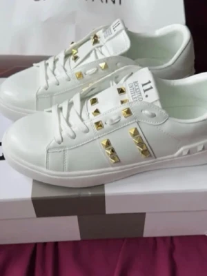 Vita sneakers med guldnitar - Snygga vita sneakers från Valentino med coola guldiga nitar på sidorna och ovandelen. Skorna har en klassisk låg modell och är gjorda i läder med vit sula och detaljer. Perfekta för dig som vill sticka ut med en lyxig touch.