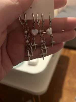 Hemma gjorda Silvriga örhängen med hjärta och stjärna - Hemma gjorda Säljer ett par trendiga silvriga örhängen med hängande kedjor, vita pärlhjärtan och metalliska stjärnberlocker. Örhängena har en cool och modern design som sticker ut. Perfekta för dig som gillar detaljer och vill ha något unikt i smyckeskrinet.