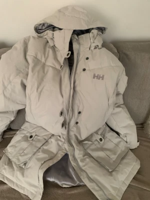 Ljusgrå dunjacka från Helly Hansen - Säljer en ljusgrå dunjacka från Helly Hansen med huva och stora fickor framtill. Jackan har dragkedja och tryckknappar, samt logga på bröstet och ryggen. Perfekt för kalla vinterdagar och riktigt varm tack vare dunfyllningen.