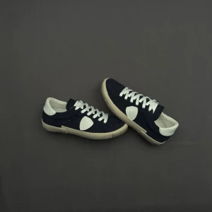 Mörkblå Philippe Model sneakers - Snygga mörkblå sneakers från Philippe Model med vita detaljer och vit sula. Skorna har klassisk låg profil, vita skosnören och är tillverkade i mocka och läder. Perfekta för en stilren och trendig look.