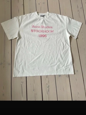 Vit Acne Studios t-shirt med tryck - Vit t-shirt från Acne Studios med rosa texttryck 'Acne Studios STOCKHOLM 1996' på bröstet. Klassisk passform med rund hals och korta ärmar. 