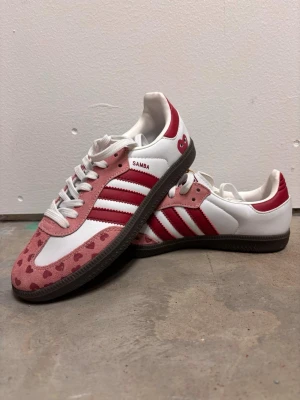 Adidas Samba rosa/röd hjärtmönster, strl 39 1/3 - Unika Adidas Samba sneakers i vitt, rosa och rött med hjärtmönster på tåpartiet. Skorna har klassiska tre ränder i rött på sidan, vit ovandel i läder och rosa mockadetaljer. Yttersulan är mörkbrun och skorna har vita skosnören.