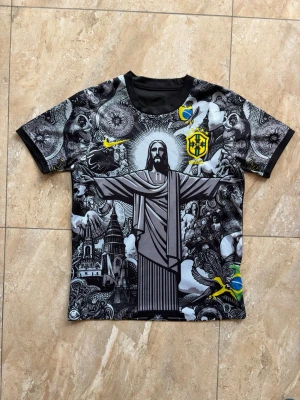 Jesus Brasilien t-shirt  - Unik svart t-shirt med stort grafiskt tryck av Kristusstatyn i Rio, detaljerade illustrationer och Brasilien-emblem på bröstet. T-shirten har rund hals, korta ärmar och inslag av gult och grönt i trycket. Perfekt för dig som gillar streetwear och fotbollsinspirerad stil.