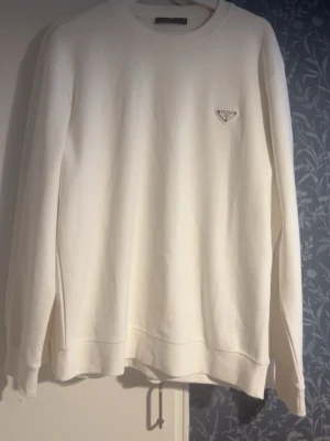 Vit sweatshirt från Prada - Snygg vit sweatshirt från Prada med klassisk rund hals och lång ärm. Tröjan har en ikonisk metallpatch med Prada-loggan på bröstet. Tillverkad i mjukt material som ger en clean och stilren look.