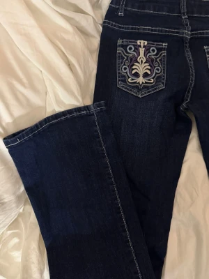 Mörkblå jeans med broderad ficka - Snygga mörkblå jeans med unik broderad bakficka i beige, lila och grått. Jeansen har raka ben och kontrastsömmar som ger en cool look. Perfekta för dig som vill sticka ut lite extra med detaljer. Materialet är klassisk denim.