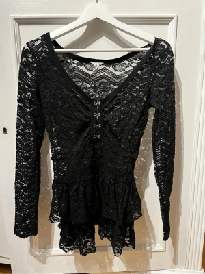 Svart spetsblus med volang -  svart blusa  spets med v-ringning och dekorativa knappar framtill. Långärmad modell med volang nedtill och transparenta detaljer. Perfekt för dig som gillar en feminin och trendig stil.