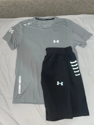 Svarta träningsshorts från Under Armour - Snygga svarta träningsshorts från Under Armour med elastisk midja och snörning. Shortsen har logga i vitt och texten 'ARMOUR' längs benet. Perfekta för gymmet eller löprundan, tillverkade i lätt och snabbtorkande material.