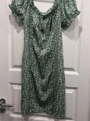 Grön blommig sommarklänning  - Söt grön sommarklänning med små vita blommor och korta puffärmar. Klänningen har en liten slits nertill och rynkdetalj vid halsringningen. Perfekt för dig som gillar romantisk och somrig stil.