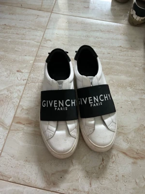 Givenchy vita sneakers med svart band - Snygga vita sneakers från Givenchy med breda svarta band över vristen där loggan står tryckt i vitt. Skorna har svarta detaljer vid hälen och Givenchy Paris-tryck i guld. Tillverkade i läder med klassisk låg profil och rundad tå.