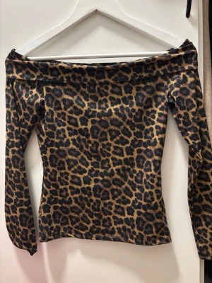 Leopardmönstrad offshoulder topp - Snygg offshoulder topp med leopardmönster i bruna och svarta toner. Toppen har långa ärmar och en tight passform som framhäver siluetten. Perfekt för dig som vill sticka ut med ett djuriskt print.