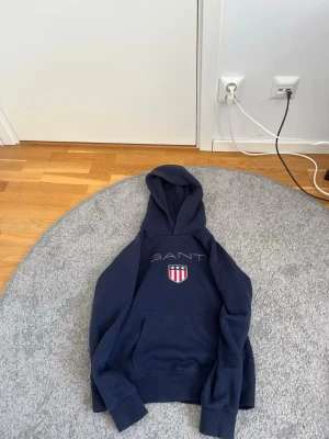 Mörkblå hoodie från GANT - Snygg mörkblå hoodie från GANT med broderad logga och ett sköldmärke på bröstet. Klassisk huva och stor magficka framtill. Perfekt för en avslappnad och sportig stil. Tillverkad i mjukt material för extra komfort.