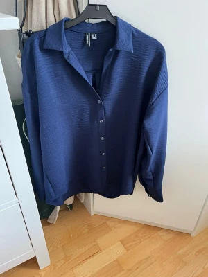 Mörkblå skjorta från Vero Moda - Snygg mörkblå skjorta från Vero Moda med klassisk krage och knappar framtill. Skjortan har lång ärm och en loose passform som ger en avslappnad vibe. Perfekt att styla med jeans eller kjol för en trendig look. Nypris 399 kr.