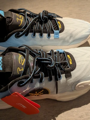 Li-Ning Way of Wade All City 12  - Snygga sneakers i ljusblått med svarta och blå snören samt svarta detaljer. Skorna har gula accenter på plösen och sidan, samt en unik design med mönstrad mesh och vadderad häl. Sulan är vit och ger en sportig känsla. Perfekta för dig som gillar streetwear.