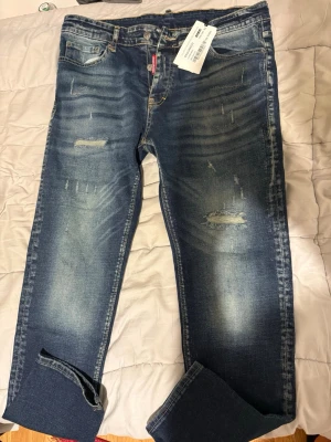 Blå slitna jeans från Dsquared2 - Säljer ett par blå jeans från Dsquared2 med slitningar och distressed detaljer. Jeansen har fem fickor, knappgylf och en snygg tvätt med ljusare partier. Perfekta för en trendig och avslappnad stil.