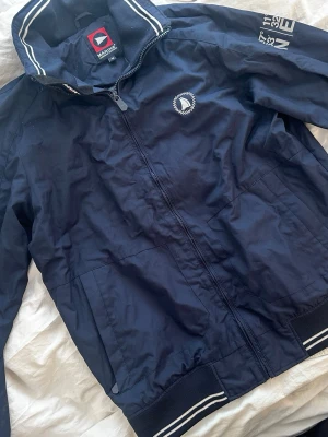 Marinblå vindjacka från Marine - Snygg marinblå vindjacka från Marine med dragkedja, två sidofickor och vita detaljer på krage och muddar. Jackan har broderad logga på bröstet och tryck på ena ärmen. Perfekt för blåsiga dagar och sportig stil.