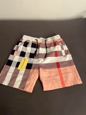 Burrbery shorts med snörning - Säljer ett par snygga burrbery shorts med klassiskt rutigt mönster i svart, vit, beige och röd. Shortsen har elastisk midja med svart snörning och sidofickor. Perfekta för sommaren och ger en trendig look till din outfit.