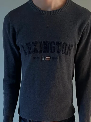 Mörkgrå stickad Lexington tröja - Mörkgrå stickad tröja från Lexington med rund halsringning och broderad logga samt texten 'Since 1993' på bröstet. Tröjan har långa ärmar och ribbade muddar vid ärmslut och nederkant. Perfekt för en avslappnad och stilren look. Skickas inom 1-3 dar vid beställning 