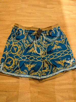 Blå mönstrade Puma shorts med snörning - Säljer ett par blå Puma shorts med guldigt barockinspirerat mönster och vita detaljer. Shortsen har snörning i midjan och elastisk linning för skön passform. Perfekta för sommaren och ger en riktigt snygg statement-look. Storlek XXXL.
