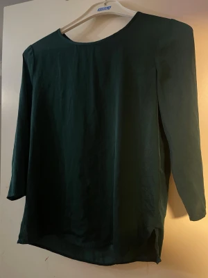 Mörkgrön blus med dragkedja bak - Enkel och stilren mörkgrön blus med rund halsringning och dragkedja i ryggen. Blusen har trekvartsärm och är tillverkad i ett lätt, slätt tyg som faller snyggt. Perfekt att matcha med jeans eller kjol för en clean look.