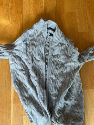 Grå kabelstickad kofta från Gant - Snygg grå kabelstickad kofta från Gant med dragkedja framtill och ribbad krage. Klassisk design med diskret logga på bröstet och ribbade muddar vid ärmslut och nederkant. Perfekt för lager på lager och enkel att matcha. Använd 1 gång bara så typ helt ny