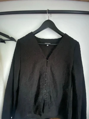 Svart blus med strukturmönster - Snygg svart blus med v-ringning och dekorativa knappar framtill. Blusen har ett unikt strukturmönster med blommiga detaljer som ger en cool touch. Långärmad och i ett mjukt tyg som känns bekvämt mot huden. Perfekt att styla med jeans eller kjol.