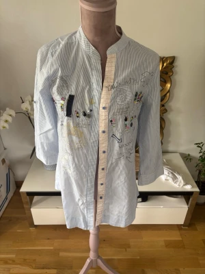 Randig skjorta med broderier och knappar från desigual - Säljer en ljusblå och vit randig skjorta med broderade detaljer och färgglada inslag framtill. Skjortan har trekvartsärm, knappar framtill och en unik broderad text på ryggen. Perfekt för dig som gillar personliga plagg med coola detaljer.