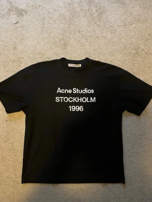 Svart Acne Studios t-shirt - Säljer en svart t-shirt från Acne Studios med vit text 'Acne Studios STOCKHOLM 1996' på bröstet. Klassisk rund hals och korta ärmar. Perfekt för dig som gillar stilrena och ikoniska plagg.