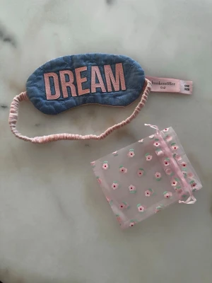 Blå sovmask / ögonbindel med text DREAM - Sovmask / ögonbindel från Hunkemöller i blått bomullstyg med rosa broderad text 'DREAM' framtill. Masken har ett mjukt, rosa elastiskt band och kommer med en söt, genomskinlig förvaringspåse med små blommor (egen). Perfekt för att blockera ljus när du ska sova. Knappt använd!