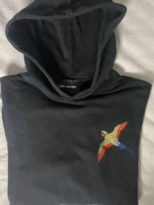 Svart hoodie med broderad fågel Axel Arigato - Säljer en svart hoodie från Axel Arigato i 100% bomull. Hoodien har en broderad färgglad fågel på bröstet och fickan, samt klassisk huva och ribbade muddar. Perfekt för dig som gillar streetwear och vill sticka ut med snygga detaljer.
