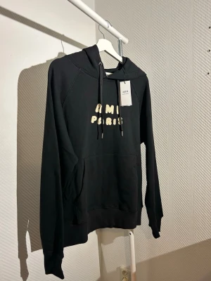 Svart hoodie från Ami Paris - Snygg svart hoodie från Ami Paris med tryckt text i beige på bröstet. Hoodien har huva med dragsko och en stor magficka framtill. Tillverkad i mjukt material som känns skönt mot huden. Perfekt för en avslappnad och trendig look.