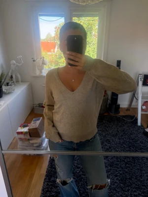 Beige stickad tröja med v-ringning - Mysig beige stickad tröja med djup v-ringning och långa ärmar. Tröjan har ribbstickade muddar vid ärmslut och nederkant, vilket ger en avslappnad men ändå stilren look. Perfekt att matcha med jeans för en enkel och snygg outfit.