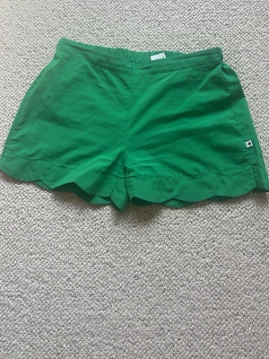 Gröna scallop shorts med resårmidja - Snygga gröna shorts med scallop-detaljer längs bensluten och bekväm resår i midjan. Perfekta för varma dagar och har en enkel, stilren look. Liten logga på sidan ger en extra touch.