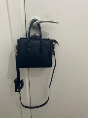 Svart minihandväska med axelrem - Snygg svart minihandväska i fyrkantig modell med både handtag och justerbar axelrem. Väskan har guldfärgade detaljer och dragkedja upptill. Perfekt för dig som gillar stilrena accessoarer och vill ha en kompakt väska till mobilen och småsaker.