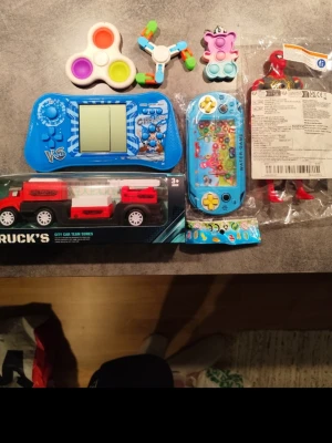 Blandat paket med leksaker - Säljer ett blandat paket med leksaker som inkluderar en röd och svart lastbilsleksak i förpackning, två blå handhållna spel, tre olika fidget-leksaker i färgglada färger samt en röd actionfigur. Perfekt för den som gillar olika typer av leksaker och spel.