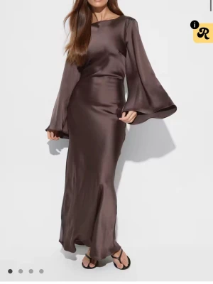 Brun långklänning i satin med vida ärmar - Helt ny i förpackning. Aldrig använd, säljer då den var fel storlek!Elegant brun långklänning i glansigt satinmaterial med vida, långa ärmar och rund halsringning. Klänningen har en figurnära passform och faller snyggt längs kroppen för en stilren look.