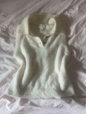 Vit stickad väst/topp med krage från H&M - Säljer en mjuk och mysig vit stickad väst/topp från H&M. Västen har en bred krage, knappar framtill och ribbade kanter. Perfekt att styla över en skjorta eller t-shirt för en trendig lager-på-lager-look.