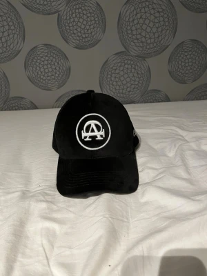 Svart All Cap One keps  - Snygg svart All Cap One keps med vit brodyr framtill och texten 'All Cap One' broderat på sidan. Det är mocka runt kepsen och den ärJusterbar baktill för perfekt passform.