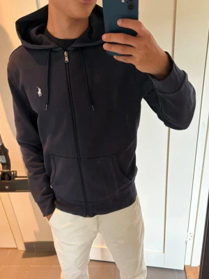Polo Ralph Lauren kofta - Mörkblå hoodie med dragkedja och huva, samt dragsnören och två fickor framtill. Liten vit logga broderad på bröstet. Perfekt för en avslappnad och stilren look.