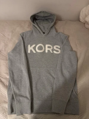 Micheal Kors grå hoodie - Snygg grå hoodie med stor vit text 'KORS' framtill. Hoodien har en klassisk huva och ribbade muddar vid ärmar och nederkant. Tillverkad i mjukt material som känns skönt mot huden. Perfekt för en avslappnad och trendig look.