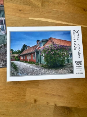 Syrenpussel 1000 bitar Gamla Gävle - Pussel med 1000 bitar från Jengel som föreställer en idyllisk gata i Gamla Gävle med röda och gröna trähus, kullerstensgata och blommande syrener. Färgstark bild i storleken 68x48 cm. Perfekt för dig som gillar utmanande pussel och vackra motiv.