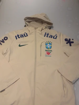 Beige Brasil vindjacka med huva - Snygg beige vindjacka med huva och dragkedja. Jackan har Brasil-logga, Nike-logga och flera sponsortryck i blått och grönt. Praktisk bröstficka med dragkedja och två sidofickor. Perfekt för dig som gillar sportig stil och vill sticka ut.