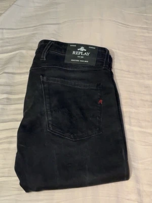 Svarta Replay anbass  - Säljer ett par svarta jeans från Replay med klassisk rak passform. Jeansen har fem fickor, bälteshällor och en liten röd detalj på bakfickan. Materialet är mjukt och stretchigt, perfekt för en avslappnad men snygg stil. Endast använda några gånger.