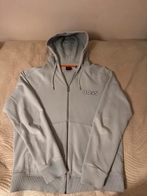 Ljusblå hoodie med dragkedja från BOSS - Snygg ljusblå hoodie från BOSS med dragkedja framtill och broderad logga på bröstet. Hoodien har snörning i huvan och två fickor framtill. Perfekt för en chill och stilren look.