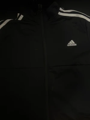 Svart Adidas träningsjacka - Snygg svart träningsjacka från Adidas med klassiska vita ränder längs ärmarna och Adidas-logga på bröstet. Jackan har dragkedja framtill och hög krage. Perfekt för träning eller chill. Materialet är troligtvis polyester.