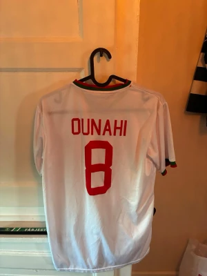 Marocko fotbollströja Puma nr 8 - Säljer en vit Marocko fotbollströja från Puma med röd text och nummer 8 (Ounahi) på ryggen. Tröjan har gröna och röda detaljer vid krage och ärmslut samt Marockos landslagsemblem på bröstet. Perfekt för match eller träning.