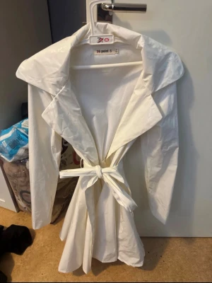 Regn jacka från fashion nova  - Snygg vit trenchcoat med bred krage och knytskärp i midjan. Jackan har långa ärmar och en klassisk siluett som ger en clean och modern look. Materialet ser ut att vara vattenavvisande, perfekt för regniga dagar.