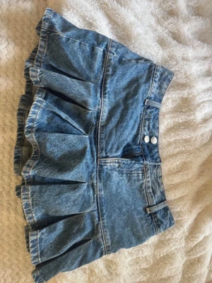 Blå jeanskjol med volang - Säljer en blå jeanskjol med volangkant och markerad söm. Kjolen har två knappar och dragkedja framtill samt hällor för skärp. Perfekt för dig som gillar en trendig och avslappnad stil. Även inbyggda shorts