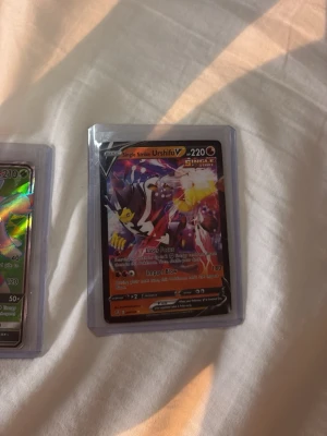 Pokémonkort Urshifu V Single Strike - Säljer ett Pokémonkort med Urshifu V Single Strike, 220 HP. Kortet har en färgstark design med röd, svart och gul bakgrund och ligger i ett plastfodral. Perfekt för samlare eller dig som vill boosta din lek med en kraftfull fighting-Pokémon.