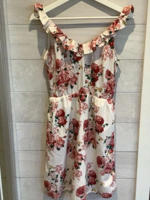 Blommig vit kortklänning med volang - Söt kortklänning i vitt med rosa och röda blommor. Klänningen har volangdetalj längs axelbanden och en lätt v-ringning. Midjan är markerad och modellen är ärmlös, perfekt för varma dagar.