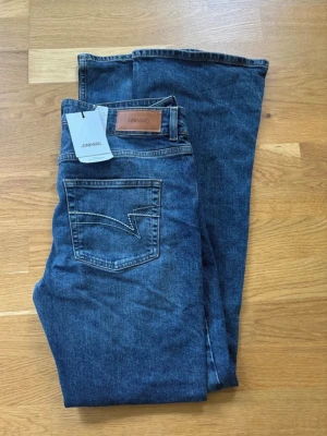 Blå bootcut jeans från Junkyard - Säljer ett par blå bootcut jeans från Junkyard med klassisk femficksdesign och snygga kontrastsömmar. Jeansen har en något utsvängd passform nedtill och är tillverkade i ett slitstarkt denimtyg. Perfekta för dig som gillar retrovibbar och vill ha en avslappnad look.